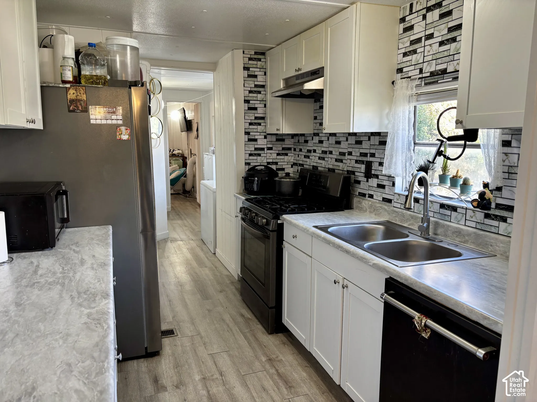 Modern kitchen featuring white cabinetry, a dual basin stainless steel sink, a black and white tiled backsplash, wood-finish flooring, and a stainless steel refrigerator