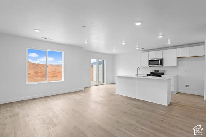Open-concept living space featuring wood-finish flooring, recessed lighting, a kitchen island with an integrated sink, white cabinetry, and stainless steel appliances