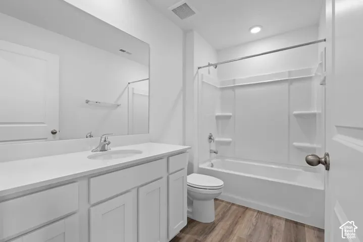 Bathroom featuring a white vanity with integrated sink, chrome faucet, a large wall-mounted mirror, white toilet, a white bathtub with shower surround, and wood-finish flooring