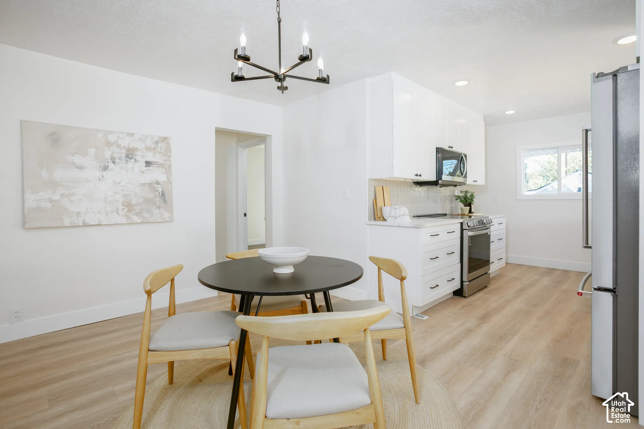 Open-concept dining and kitchen area featuring a round black table with wood-finish base, natural wood-finish flooring throughout, white cabinetry with integrated appliances, a modern pendant light fixture, and abundant natural light from multiple windows