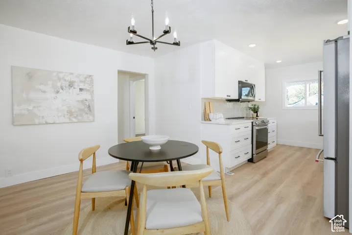 Open-concept dining and kitchen area featuring a round black table with wood-finish base, natural wood-finish flooring throughout, white cabinetry with integrated appliances, a modern pendant light fixture, and abundant natural light from multiple windows