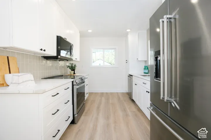 White cabinetry with matte black hardware, stainless steel appliances, light wood-finish flooring, white countertops, and a subway tile backsplash