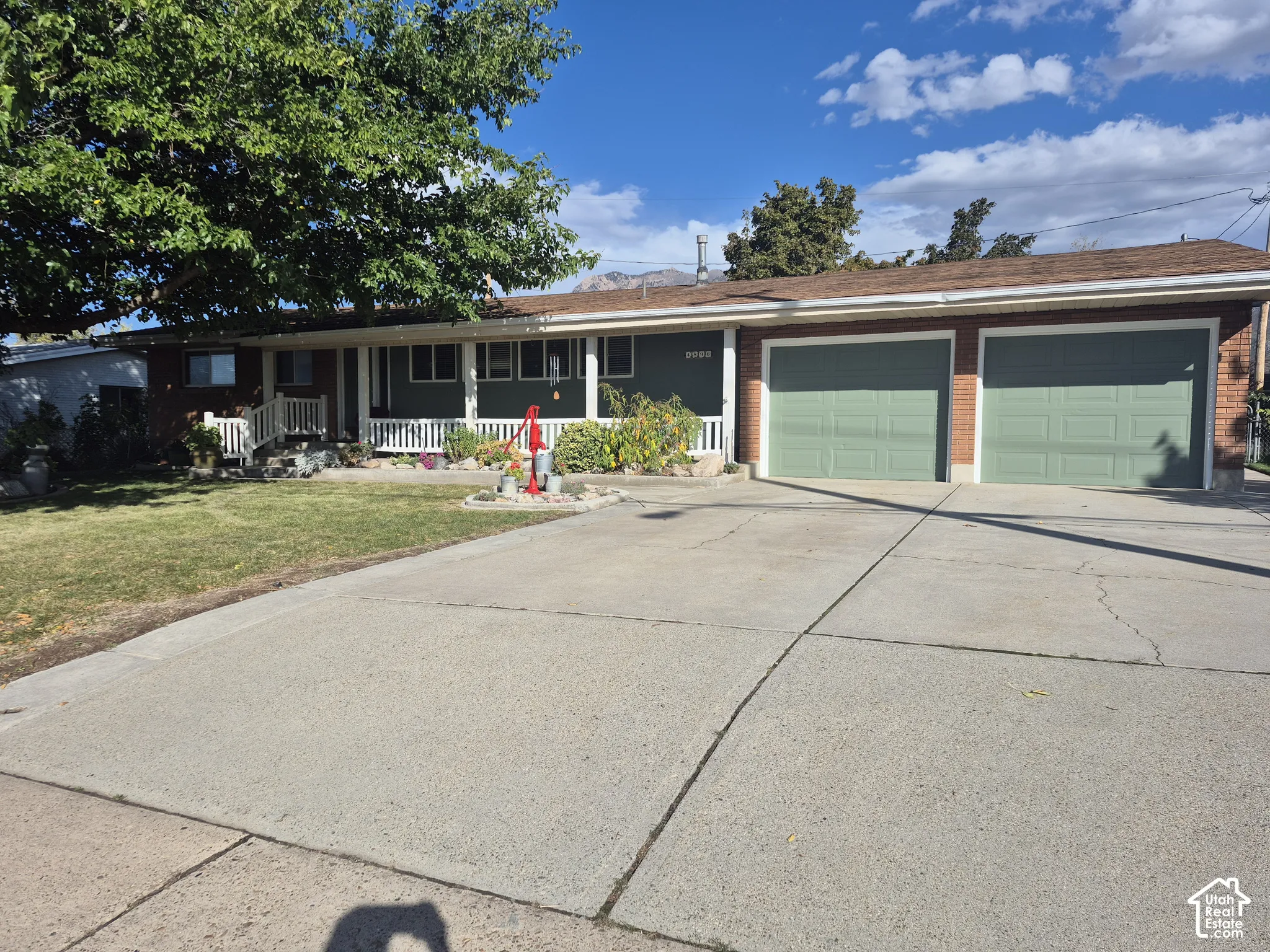 Two-car garage with light green doors, concrete driveway, brick exterior accents, covered front porch, and mature front yard landscaping