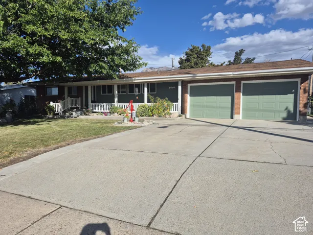 Two-car garage with light green doors, concrete driveway, brick exterior accents, covered front porch, and mature front yard landscaping