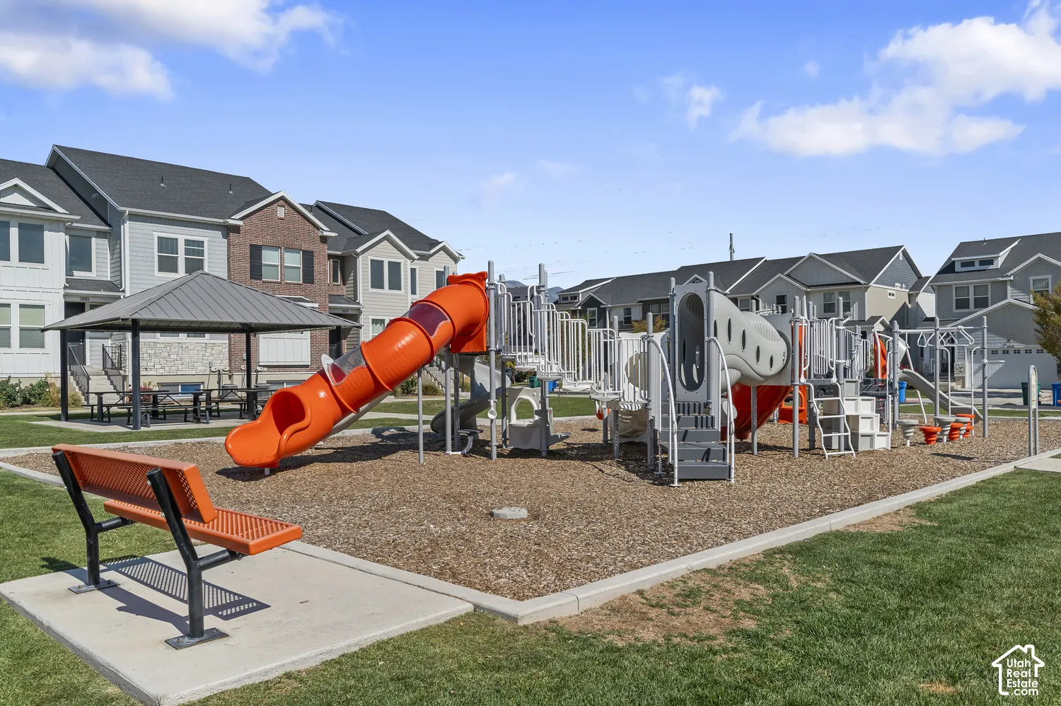 Community playground featuring a multi-level play structure with an orange tube slide, climbing apparatus, and ground-level activities, set on a mulch surface with adjacent bench seating