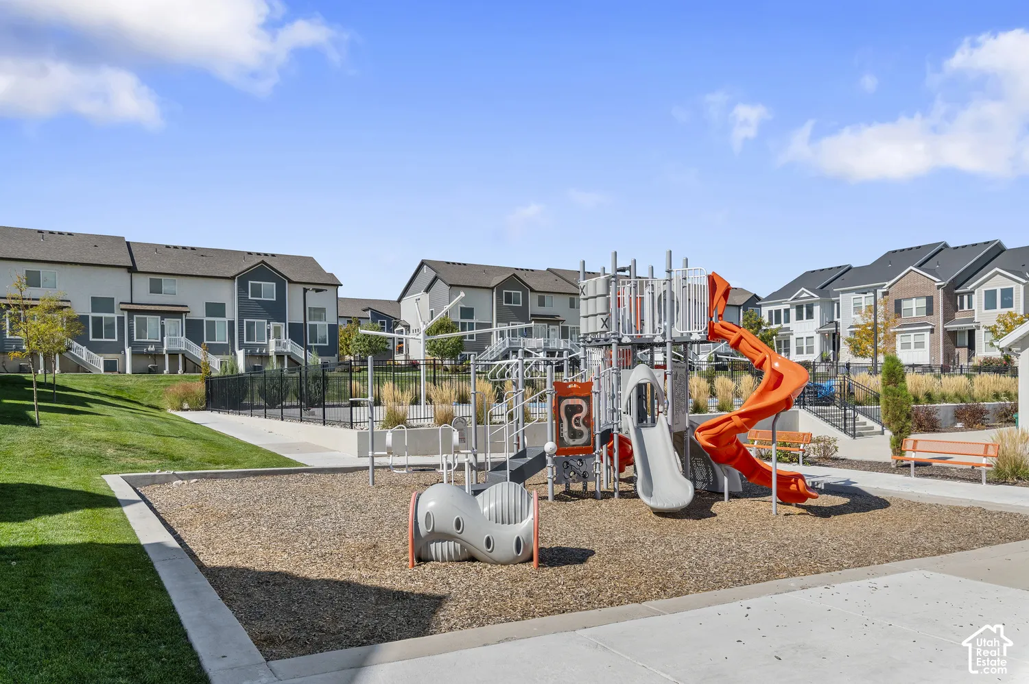 Community playground featuring modern multi-level play structures with orange accents, ground-level safety surfacing, and open sightlines