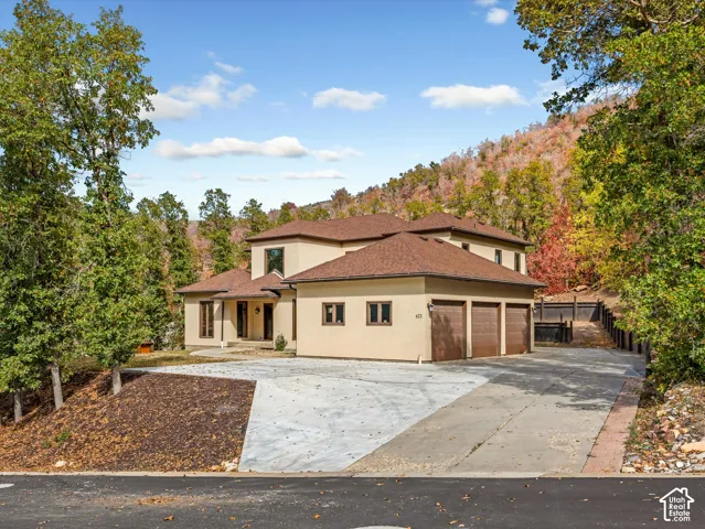 Stucco exterior featuring a multi-level roofline, three-car garage, concrete driveway, and a natural hillside backdrop