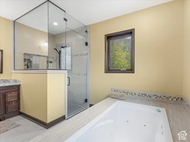 Spacious bathroom featuring a glass-enclosed shower, built-in soaking tub, wood-finish flooring, a vanity with a stone countertop, and a window providing natural light