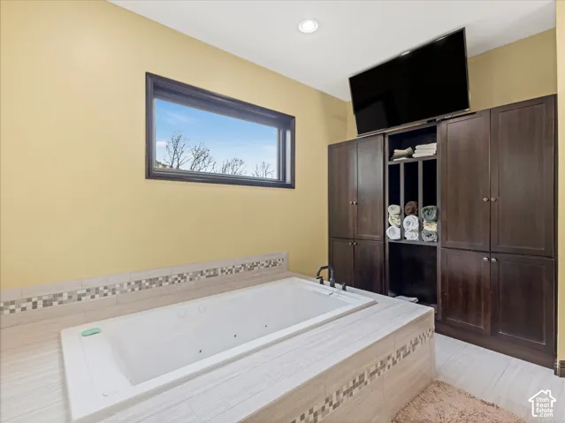 Jetted bathtub with tiled surround, wood-finish flooring, built-in cabinetry with open shelving, and a horizontal window