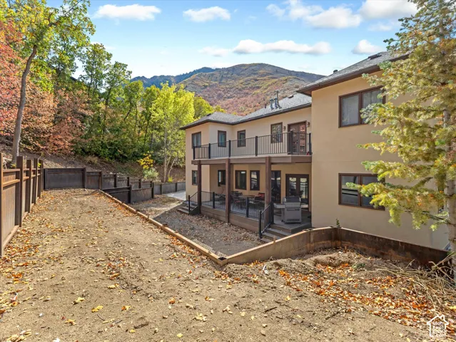 Stucco exterior residence featuring an elevated deck with black metal railings, a covered lower-level patio, terraced timber retaining walls, and a mountain backdrop
