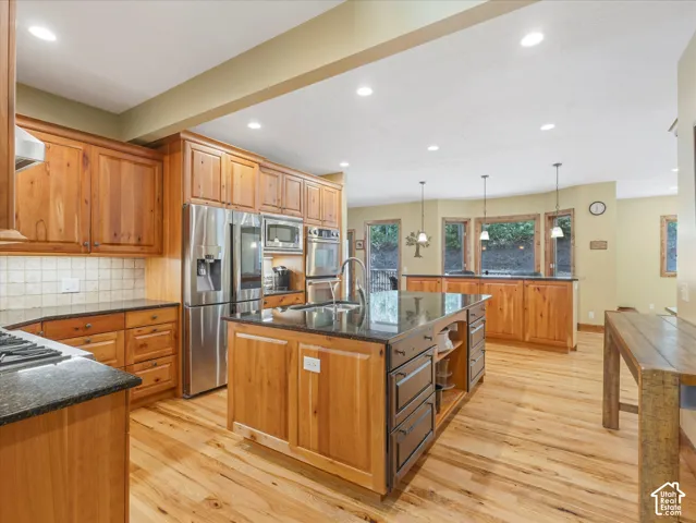Kitchen featuring natural wood cabinetry, stainless steel appliances, granite countertops, a central island with a sink, and light wood-finish flooring