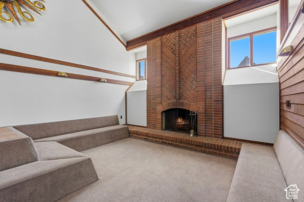 Brick fireplace with hearth, vaulted ceiling, wood trim accents, and carpeted flooring