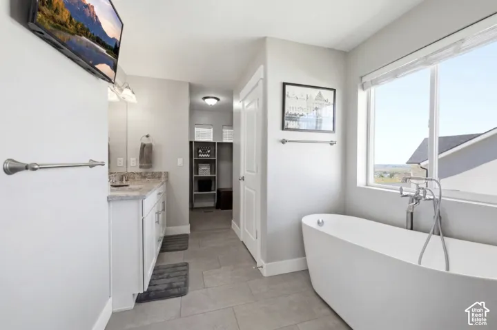 Freestanding soaking tub with floor-mounted faucet, large window, double vanity with stone countertop, and tile flooring