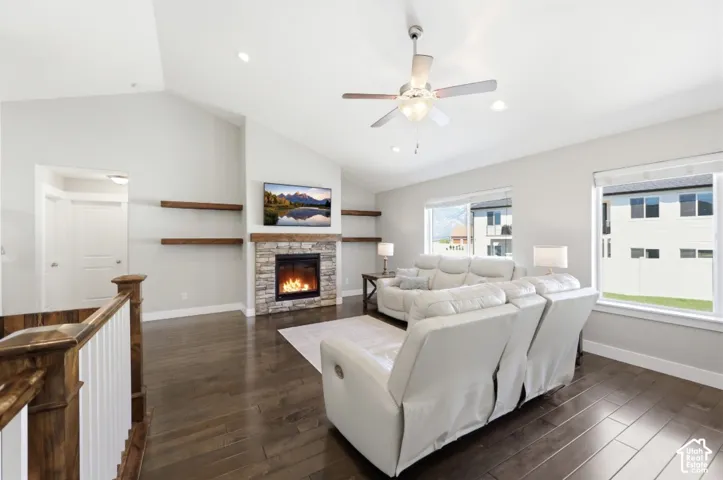 Vauted ceiling with recessed lighting and a ceiling fan, stone-clad fireplace with wood-finish mantel and floating shelves, dark wood-finish flooring, and large windows