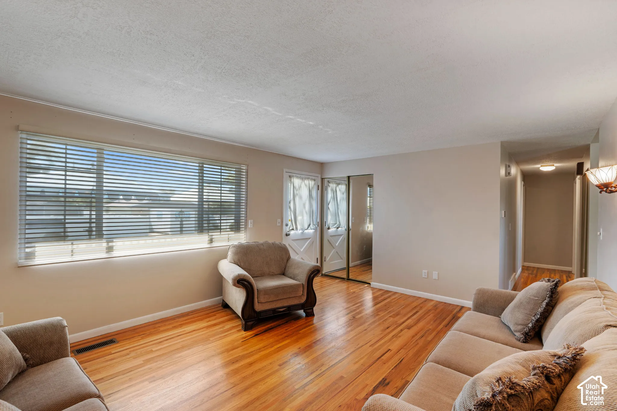 Hardwood flooring throughout, large window with blinds, dual mirrored closet doors, and a hallway with overhead lighting