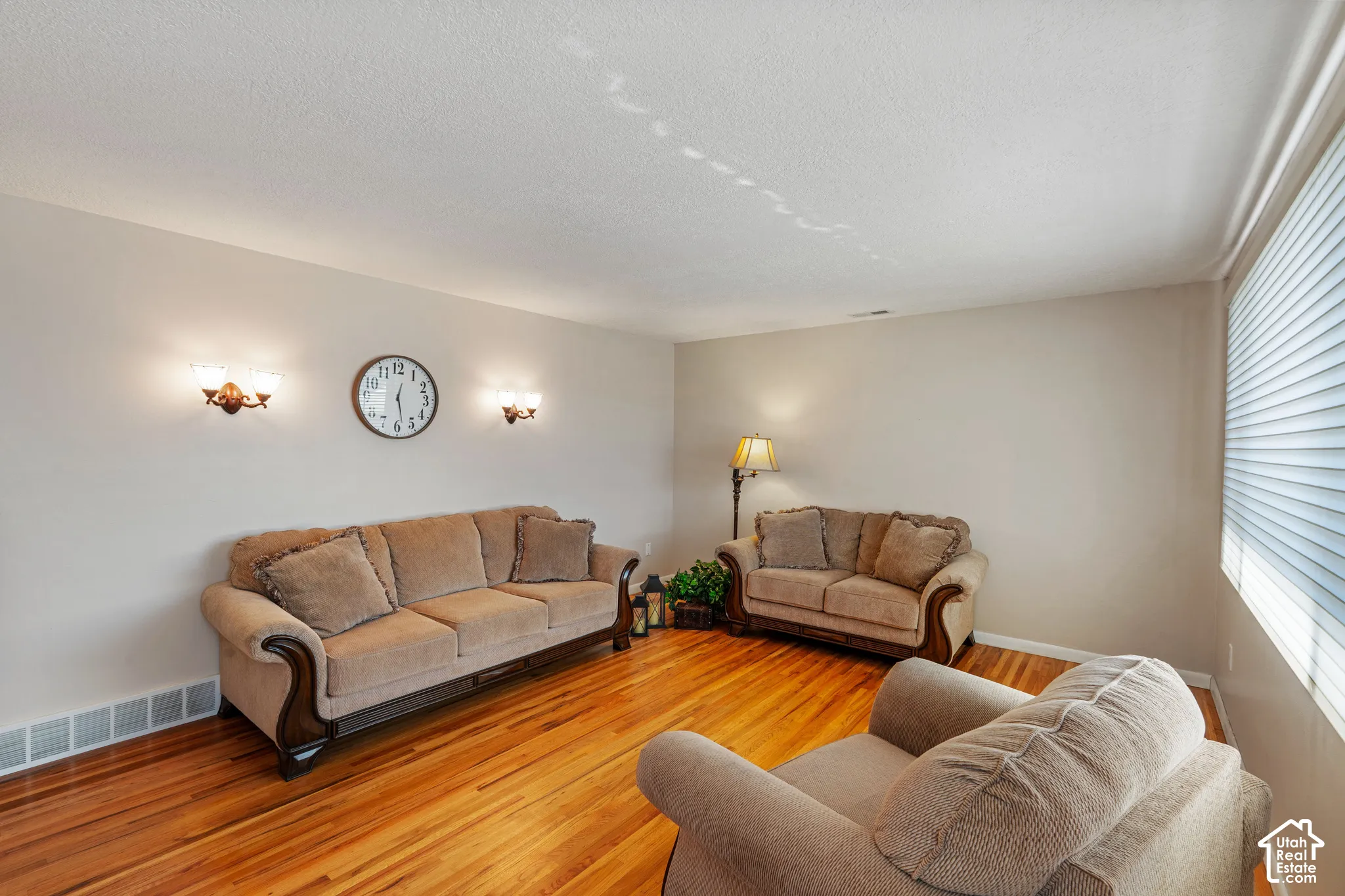 Spacious living room featuring warm wood-finish flooring, elegant upholstered seating arrangements, crown molding, recessed ceiling lighting, and wall-mounted sconces for ambient illumination