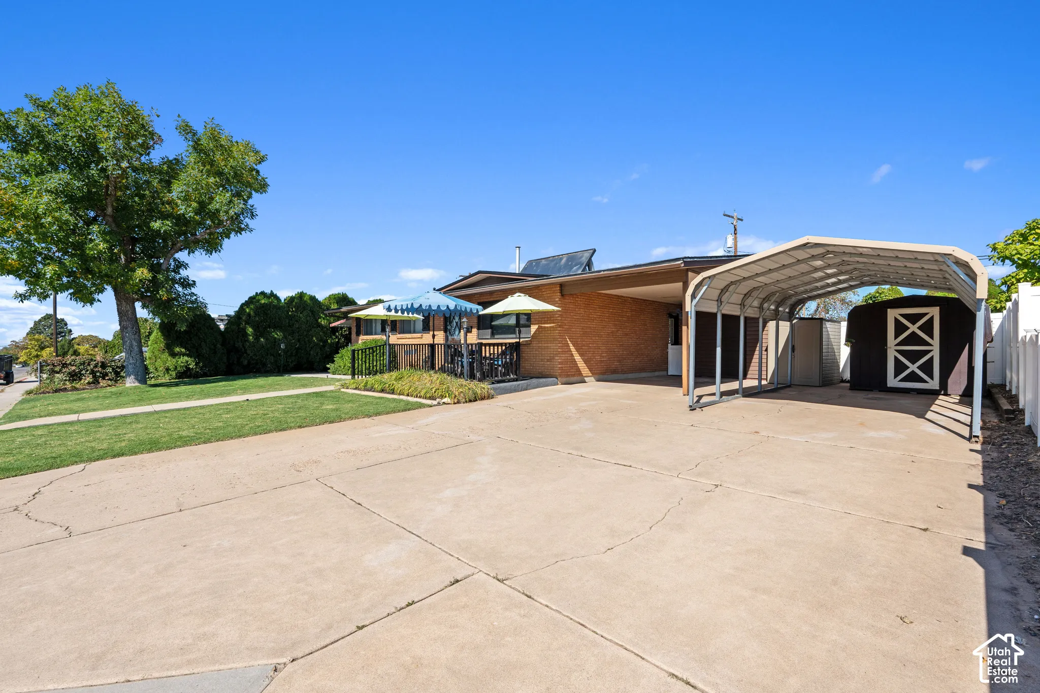 Expansive concrete driveway with a metal carport structure, brick exterior, and a detached shed