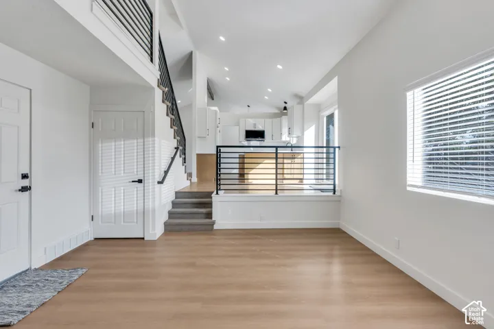 Open-concept living area featuring wood-finish flooring, a contemporary black metal stair railing, vaulted ceiling with recessed lighting, and a kitchen with white cabinetry