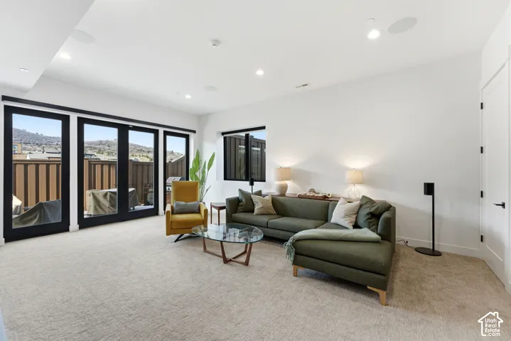 Spacious primary living area featuring floor-to-ceiling multi-panel glass doors with black frames opening to a private balcony, neutral carpeting throughout, recessed ceiling lighting, and contemporary furnishings positioned to maximize the room's generou