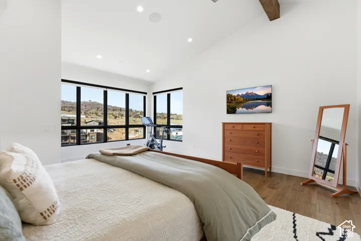 Bright primary bedroom featuring expansive black-framed windows with panoramic mountain vistas, soaring vaulted ceilings with exposed wood beams, recessed lighting, wood-finish flooring, and a substantial walk-in closet