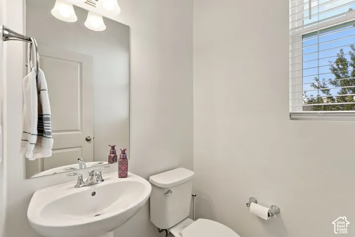 Pedestal sink with chrome faucet, wall-mounted mirror, overhead vanity lighting, white window blinds, and a white toilet