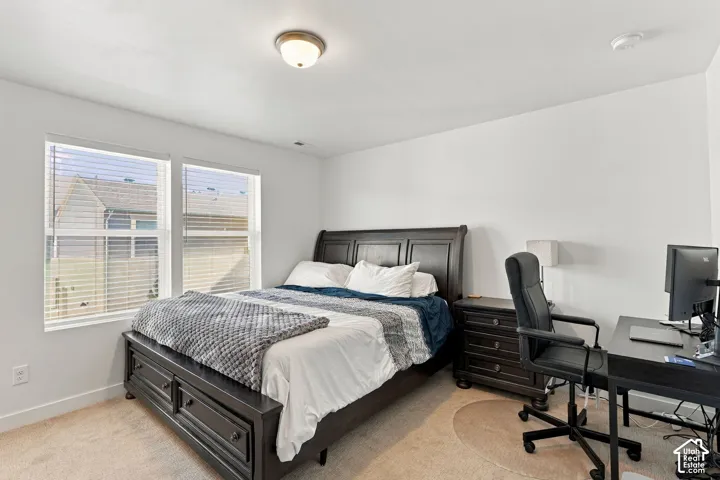 Spacious bedroom with abundant natural light from dual windows, dark wood-finish bed frame with storage drawers, neutral carpeting, and dedicated workspace with desk and office chair