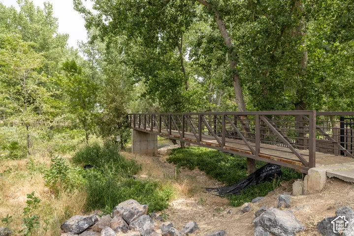 Pedestrian bridge with metal railings and a concrete support structure, surrounded by mature trees and natural landscaping