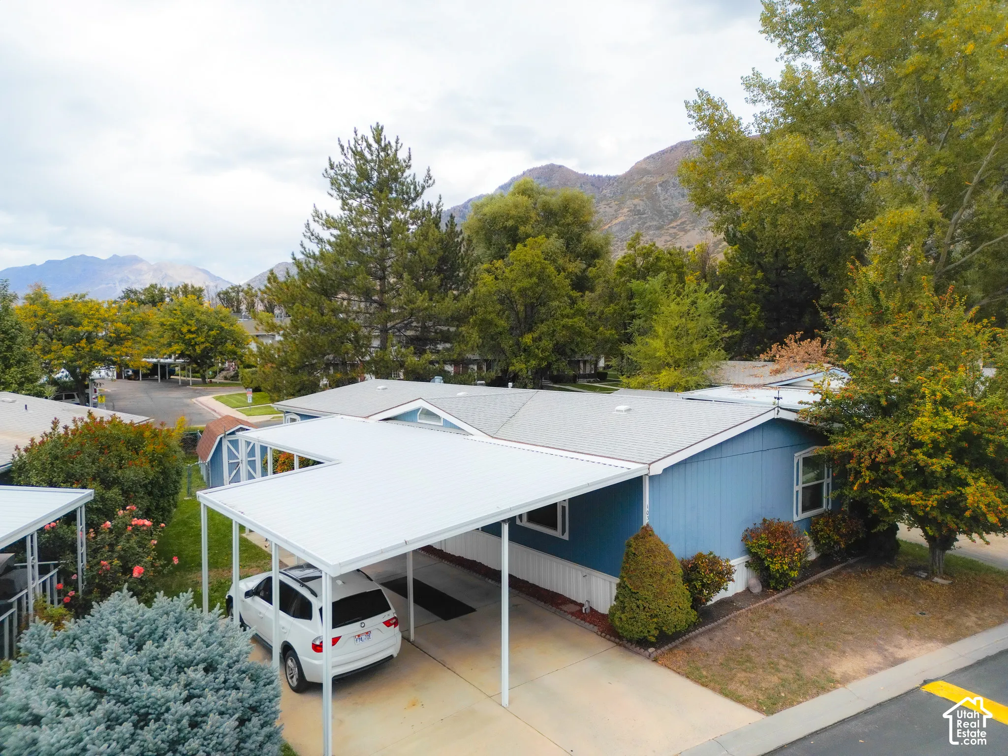 Exterior featuring a covered carport, blue siding, a grey shingle roof, mature trees, and surrounding mountain scenery