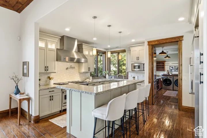 Kitchen featuring wood-finish flooring, an oversized island with pendant lighting, a stainless steel range hood, a tile backsplash, and a built-in microwave
