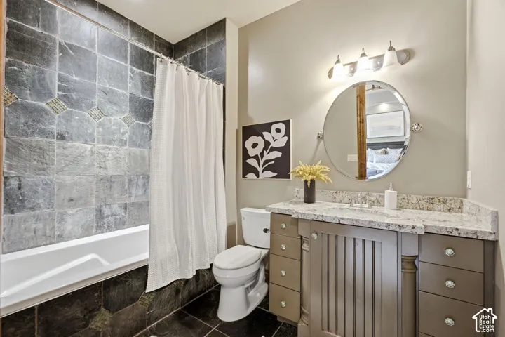 Tiled shower surround with decorative accent band, built-in bathtub, vanity with stone-finish countertop, single basin sink, and three-light vanity fixture