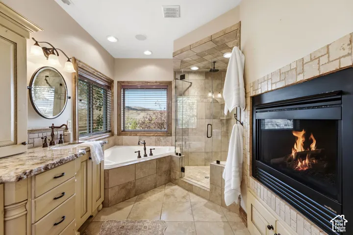 Corner soaking tub, tiled shower enclosure with glass panels, fireplace with stone surround, dual-sink vanity with stone countertop, and wood-framed windows
