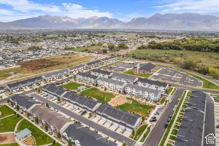 Expansive residential development featuring multi-story townhomes, manicured green spaces, paved roadways, and a prominent community building with ample parking, all set against a backdrop of distant mountains