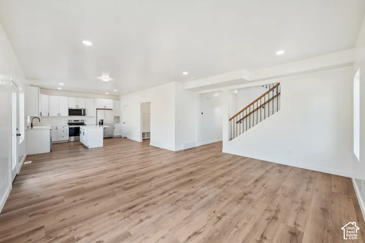 Expansive living area with wood-finish flooring, recessed lighting, a modern kitchen with white cabinetry, and a staircase featuring a wood handrail and black iron spindles