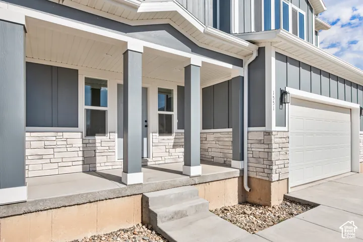 Covered concrete porch with white-framed windows, stone veneer accents, vertical siding, and an attached garage