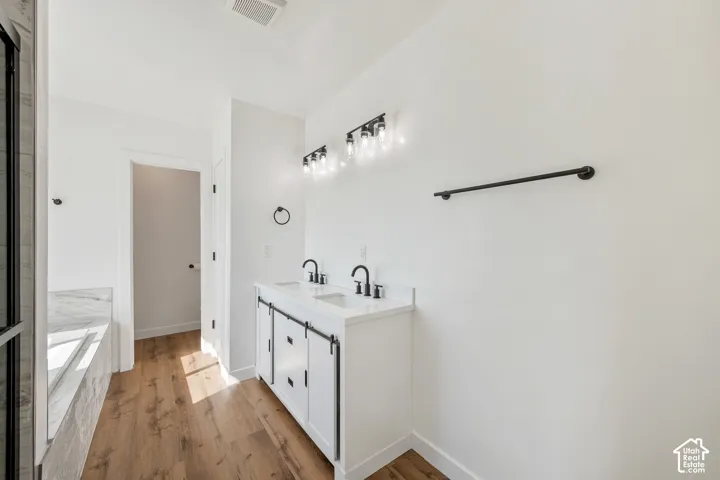 Double vanity with white cabinetry, black hardware, wood-finish flooring, and a built-in tub with marble-look surfacing