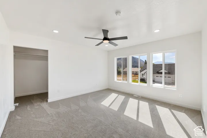 Recessed ceiling lighting, ceiling fan, three large picture windows, soft gray carpeting, and a built-in closet with hanging rod