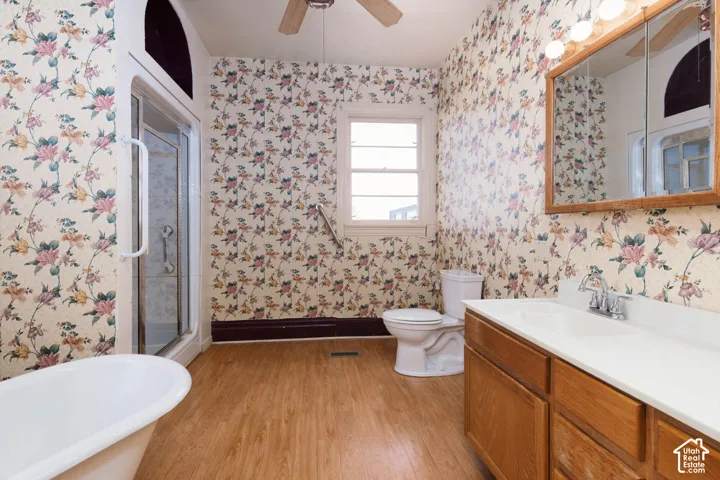 Freestanding bathtub, glass-enclosed shower, wood-finish flooring, single vanity with wood cabinetry, and a ceiling fan