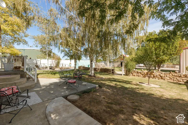 Concrete patio with a curved edge, steps with white railing, and a spacious grassy yard featuring mature trees and a stone retaining wall