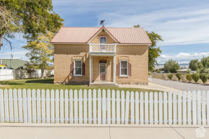 Brick exterior with a metal roof, featuring a second-story balcony, white picket fence, and a gravel driveway