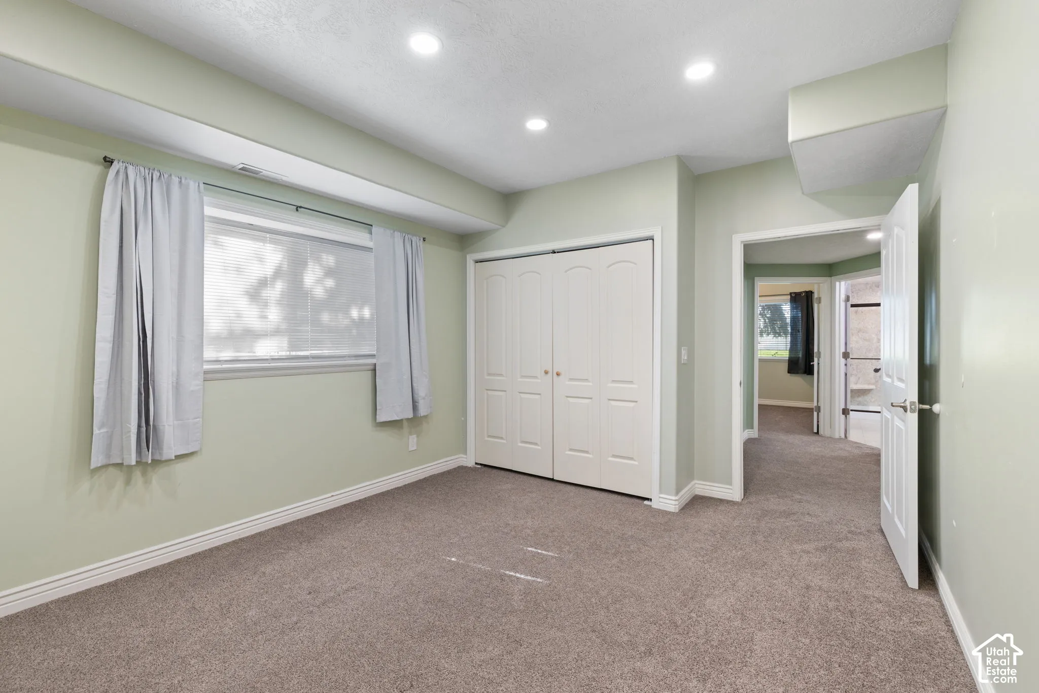 Spacious primary bedroom featuring soft sage-green walls, recessed ceiling lighting, dual closets with white panel doors, carpeting throughout, and abundant natural light from a large window with light filtering curtains