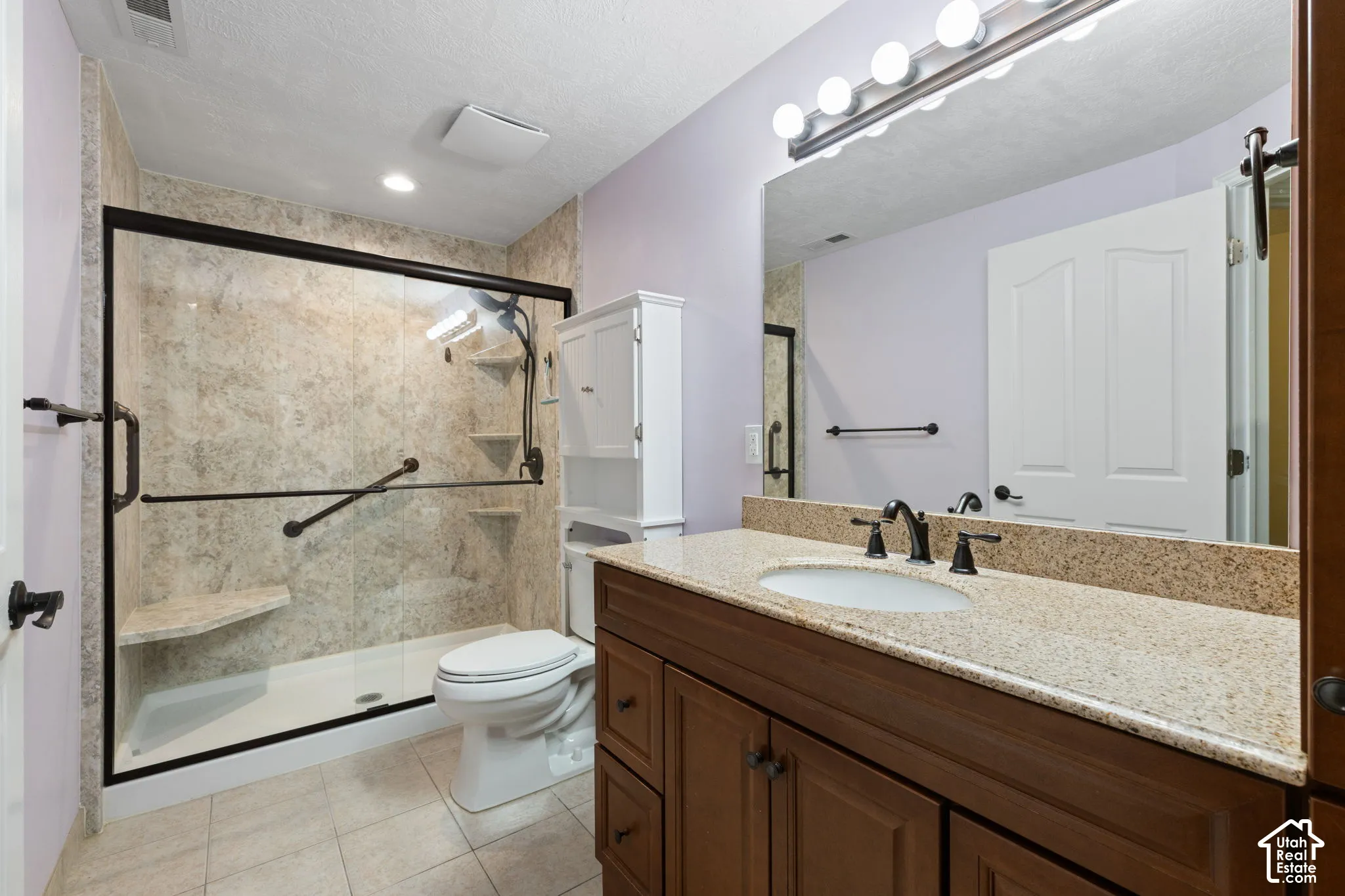 Walk-in shower with grab bars and built-in bench, wood-finish vanity with granite countertop, large vanity mirror, and tile flooring