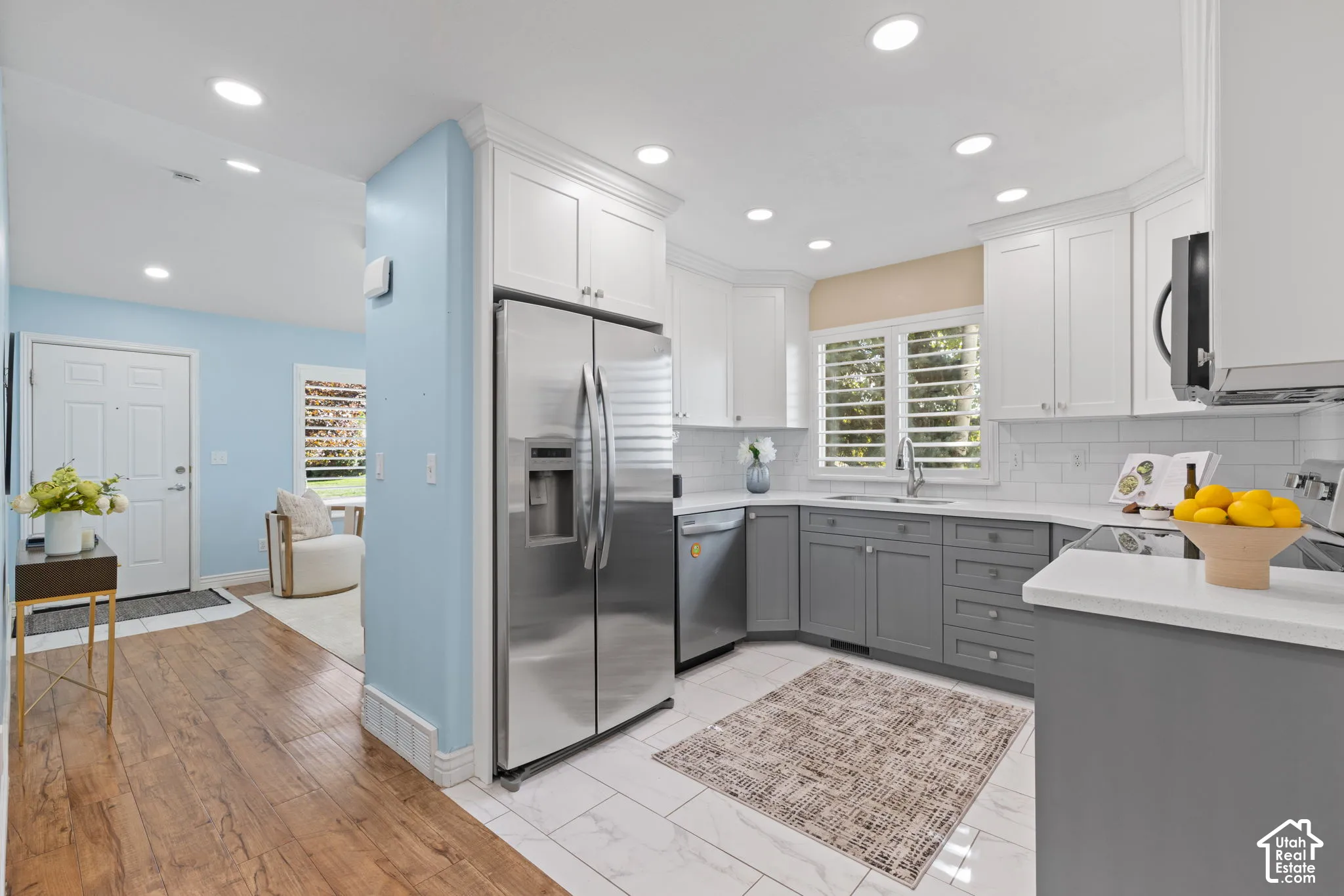 Modern kitchen featuring stainless steel side-by-side refrigerator, white cabinetry with gray lower cabinets, white subway tile backsplash, recessed ceiling lighting, and wood-finish flooring throughout the open layout