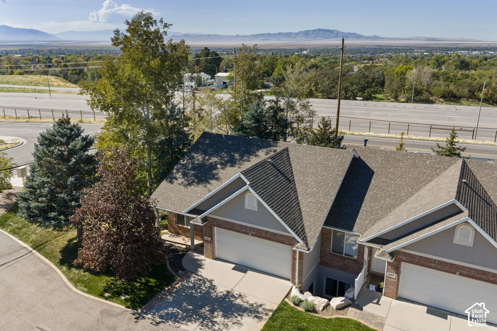Brick exterior residence featuring a multi-gabled roofline with architectural shingles, two-car garage with paneled doors, and mature landscaping including evergreen and deciduous trees