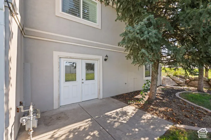 Exterior stucco finish with white trim, double glass-paneled doors, concrete patio, and established evergreen trees