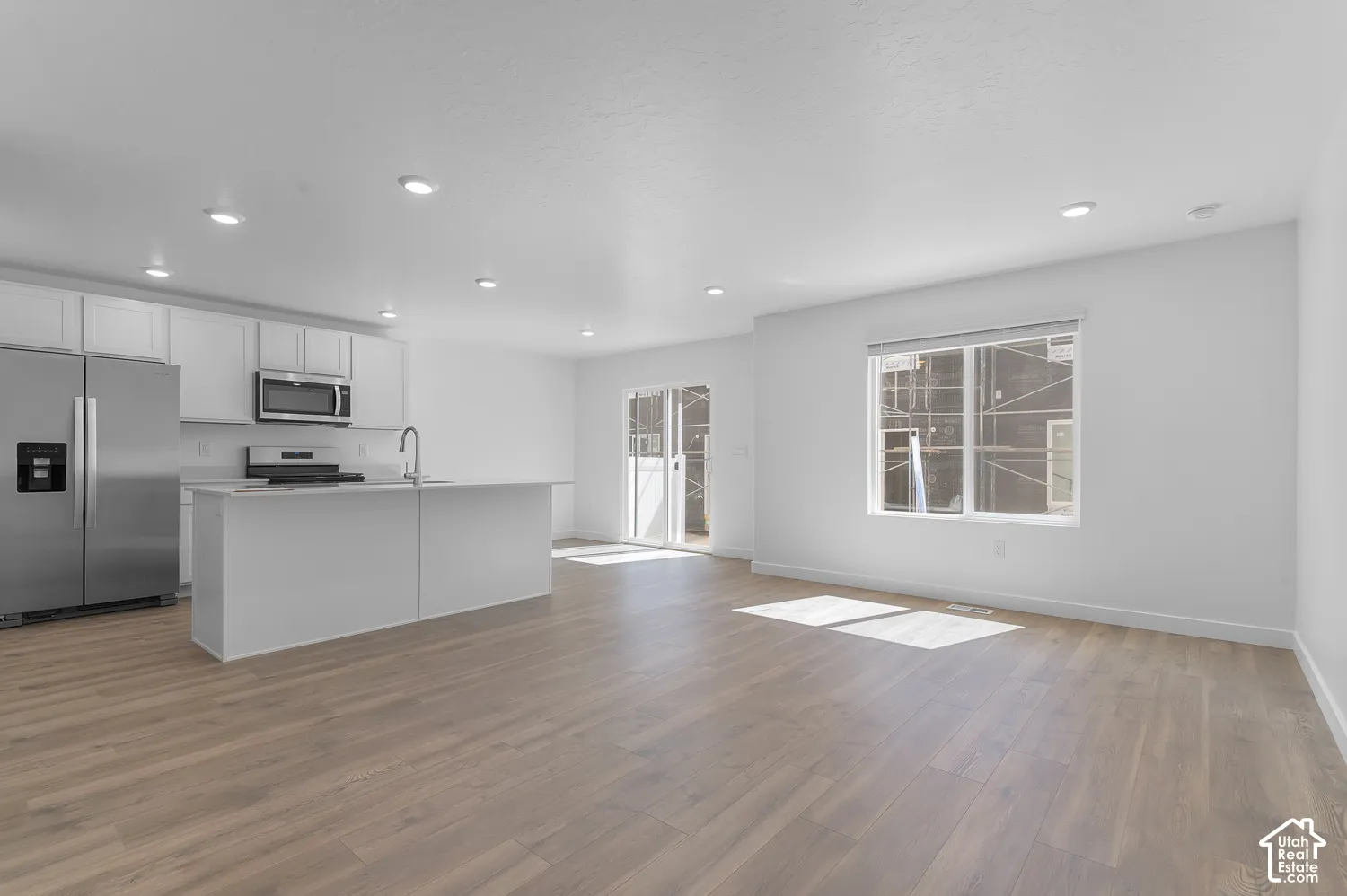 Open-concept living space with wood-finish flooring, recessed lighting, and a kitchen featuring stainless steel appliances, a center island with sink, and white cabinetry
