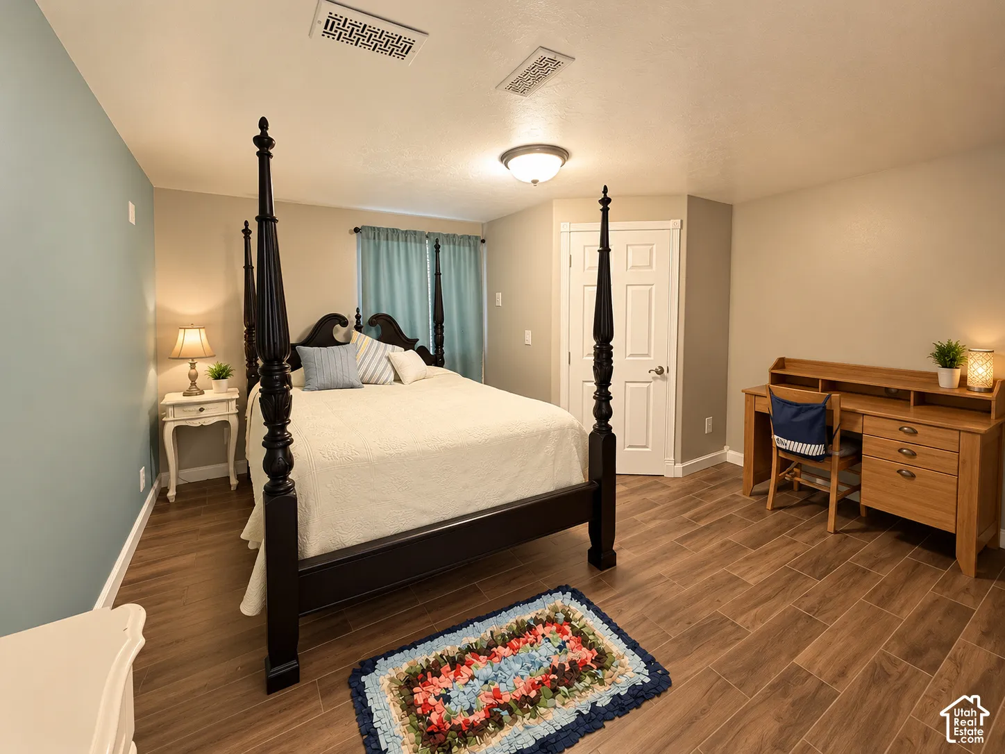 Spacious primary bedroom featuring a four-poster bed frame, wood-finish flooring throughout, soft blue accent wall, dual nightstands with task lighting, and an en-suite bathroom