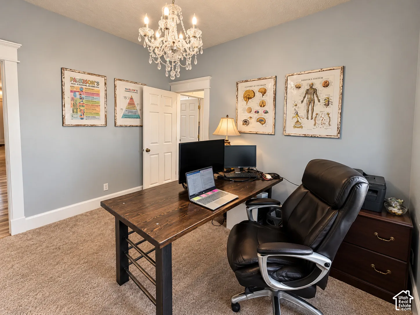 Neutral-toned wall paint, decorative chandelier light fixture, wood-finish desk with black metal frame, and light brown carpet flooring