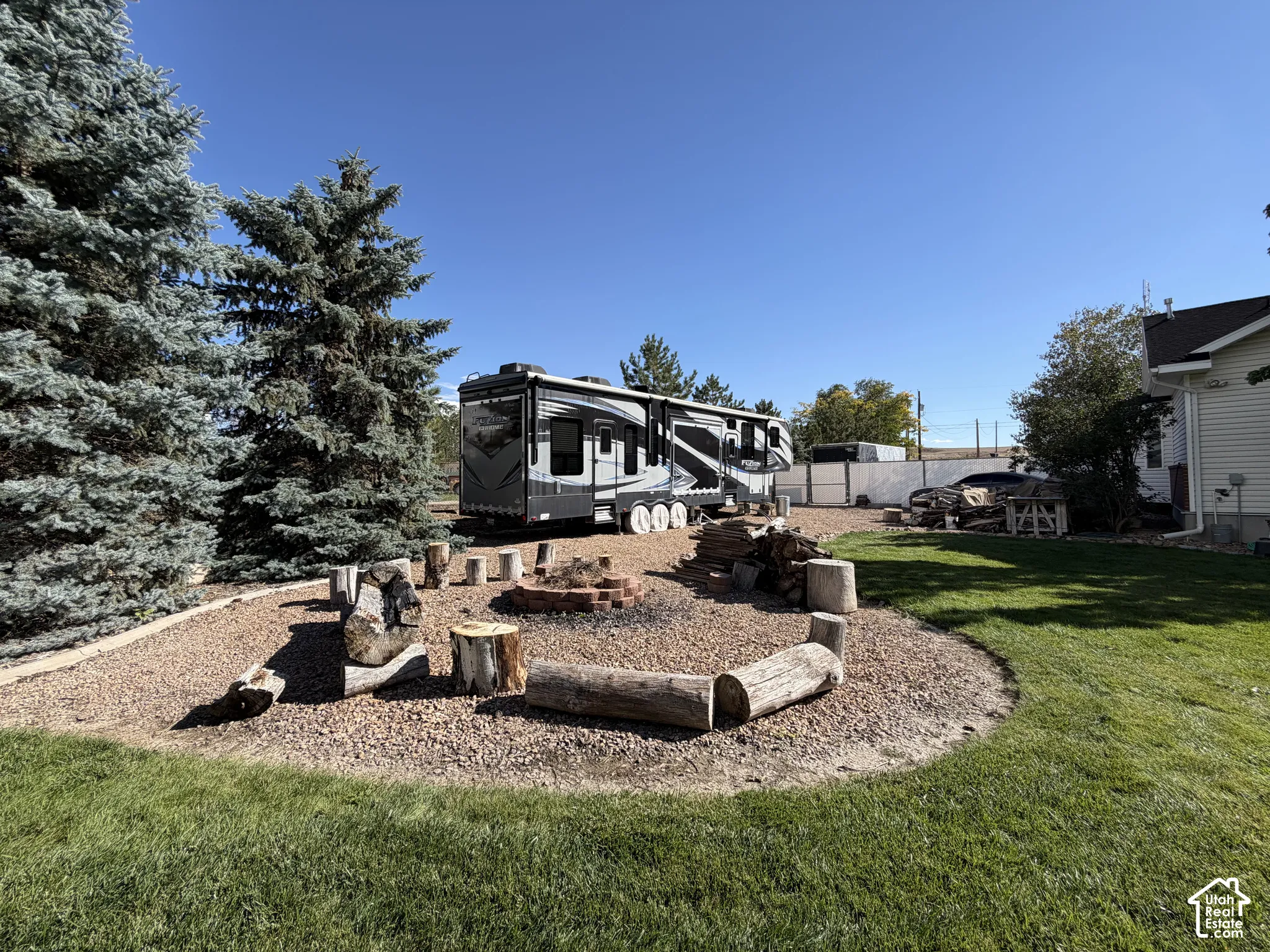 Spacious backyard retreat featuring a full-service RV pad with concrete foundation, mature landscaping including evergreen trees, decorative rock hardscaping with natural wood seating elements, manicured lawn, and detached outbuildings