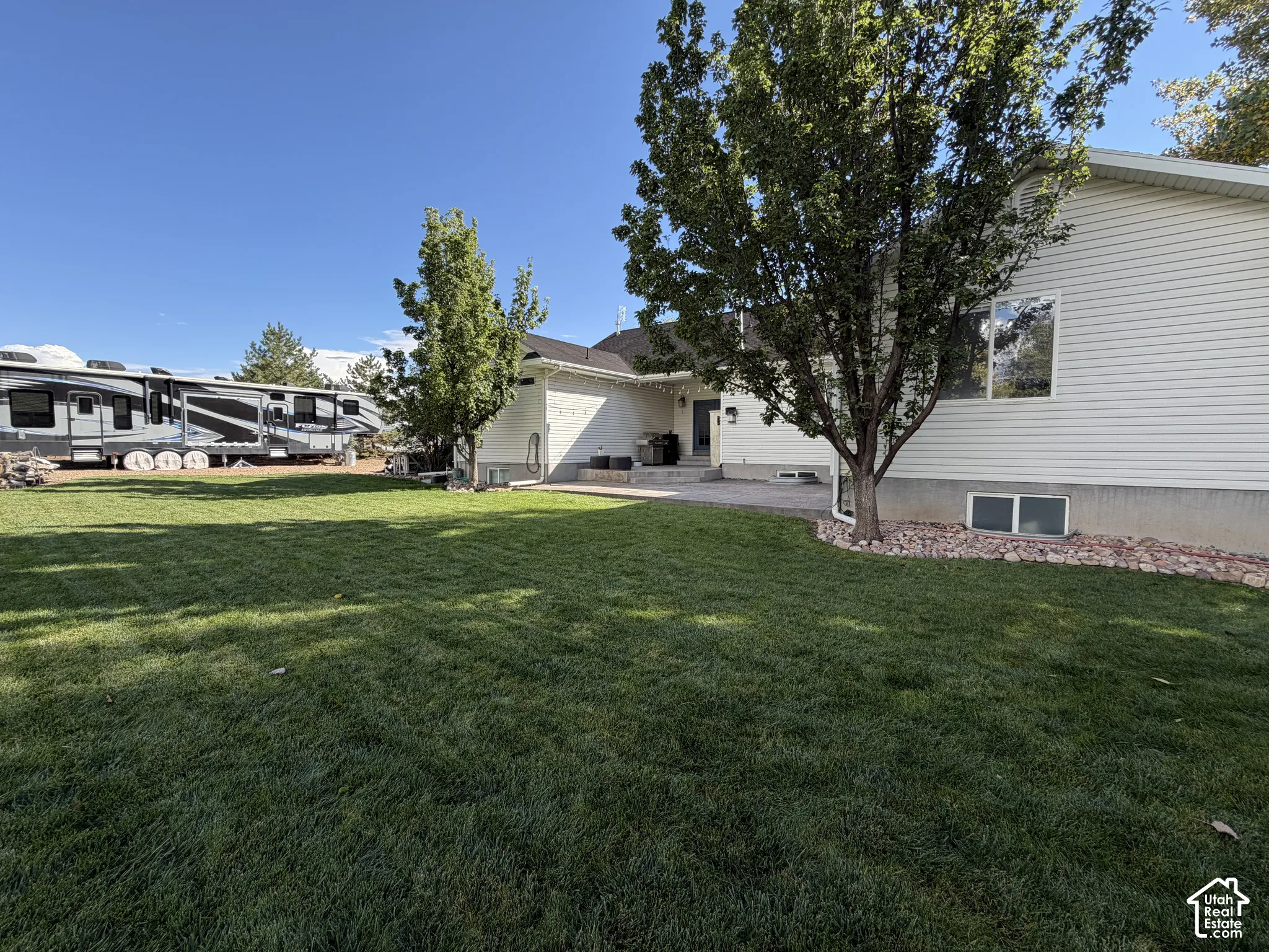 Expansive lawn area with mature trees, a paved patio, and white vinyl siding