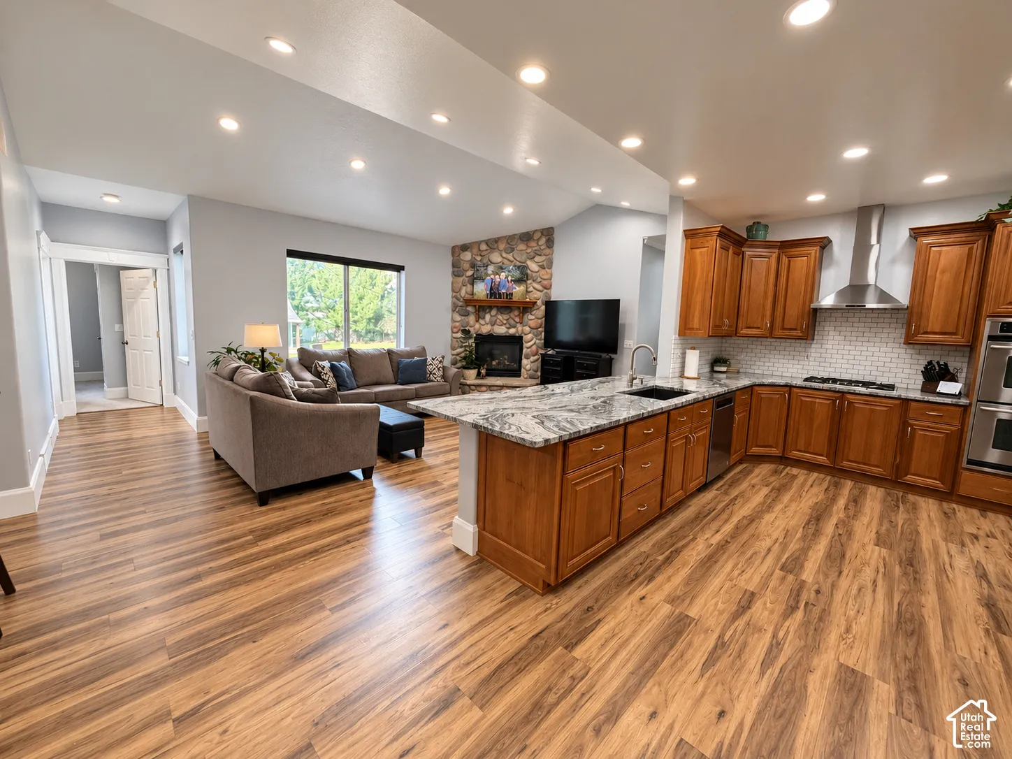Open-concept living space featuring wood-finish flooring, recessed lighting, a stone-front fireplace, a large window, and a kitchen island with stone countertops